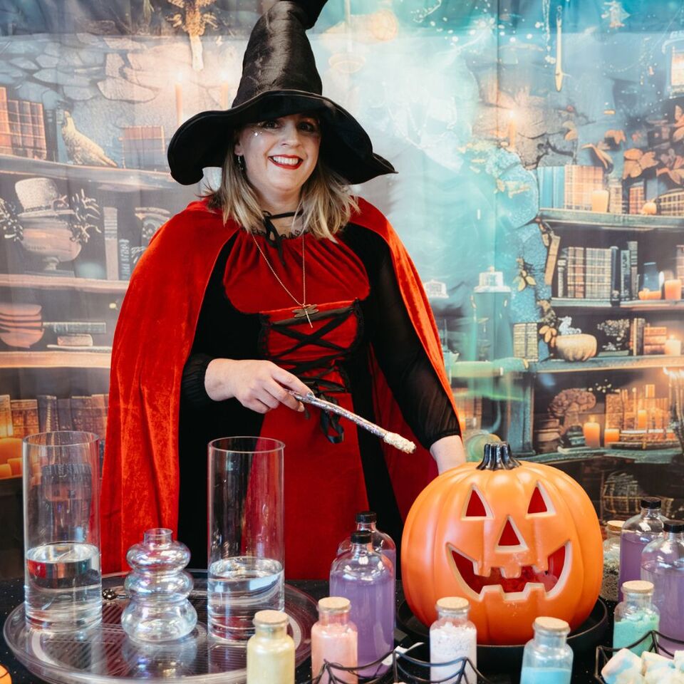 Glenda Goodspell, the Good Witch from Sparkle & Spell Co., charms the room with her vibrant red cape, sparkling wand, and potion-filled table, accompanied by a glowing pumpkin and a magical backdrop of books and candles